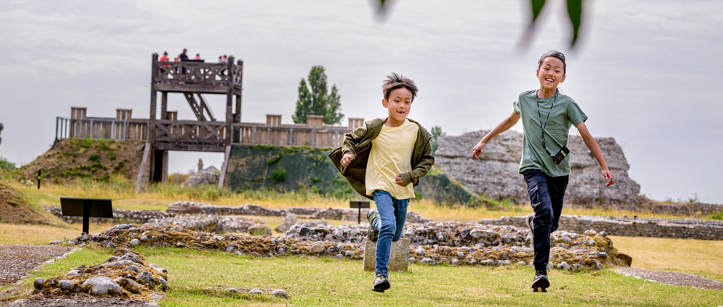 Two children run while smiling with Richborough Roman Fort ruins in the background.