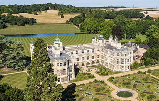 Aerial view of a grand mansion with formal gardens in front and behind a sweeping lawn and river