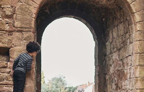 A young boy peers behind a stone archway with view of the gardens at Kenilworth Castle.