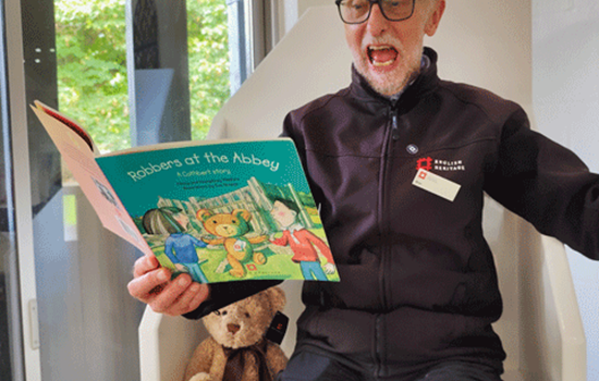 A man sits on a white chair and enthusaistically reads a children's book titled 'Robbers at the Abbey' with his arm raised in motion. He is wearing English Heritage uniform.